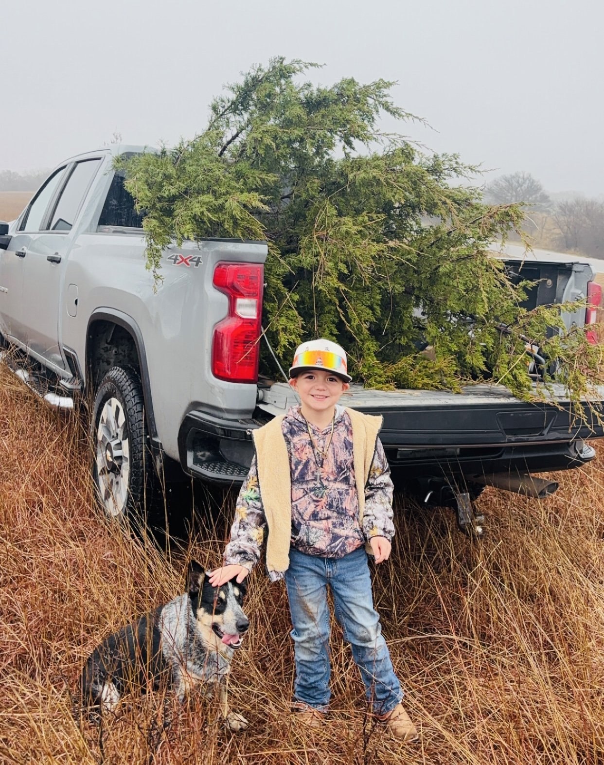 A young boy wearing a colorful hat and camouflage clothing stands beside a black and white dog in a grassy field, with a pickup truck in the background loaded with branches. The scene captures a day of outdoor adventure and bonding with pets.