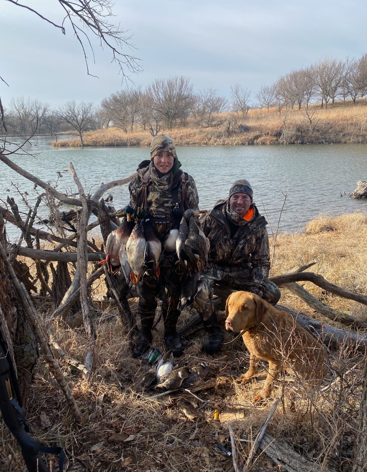 Two hunters in camouflage gear proudly display their catch of ducks by a lakeside, accompanied by a hunting dog. The scene captures a winter day with bare trees and dry grass, highlighting the outdoor hunting experience.