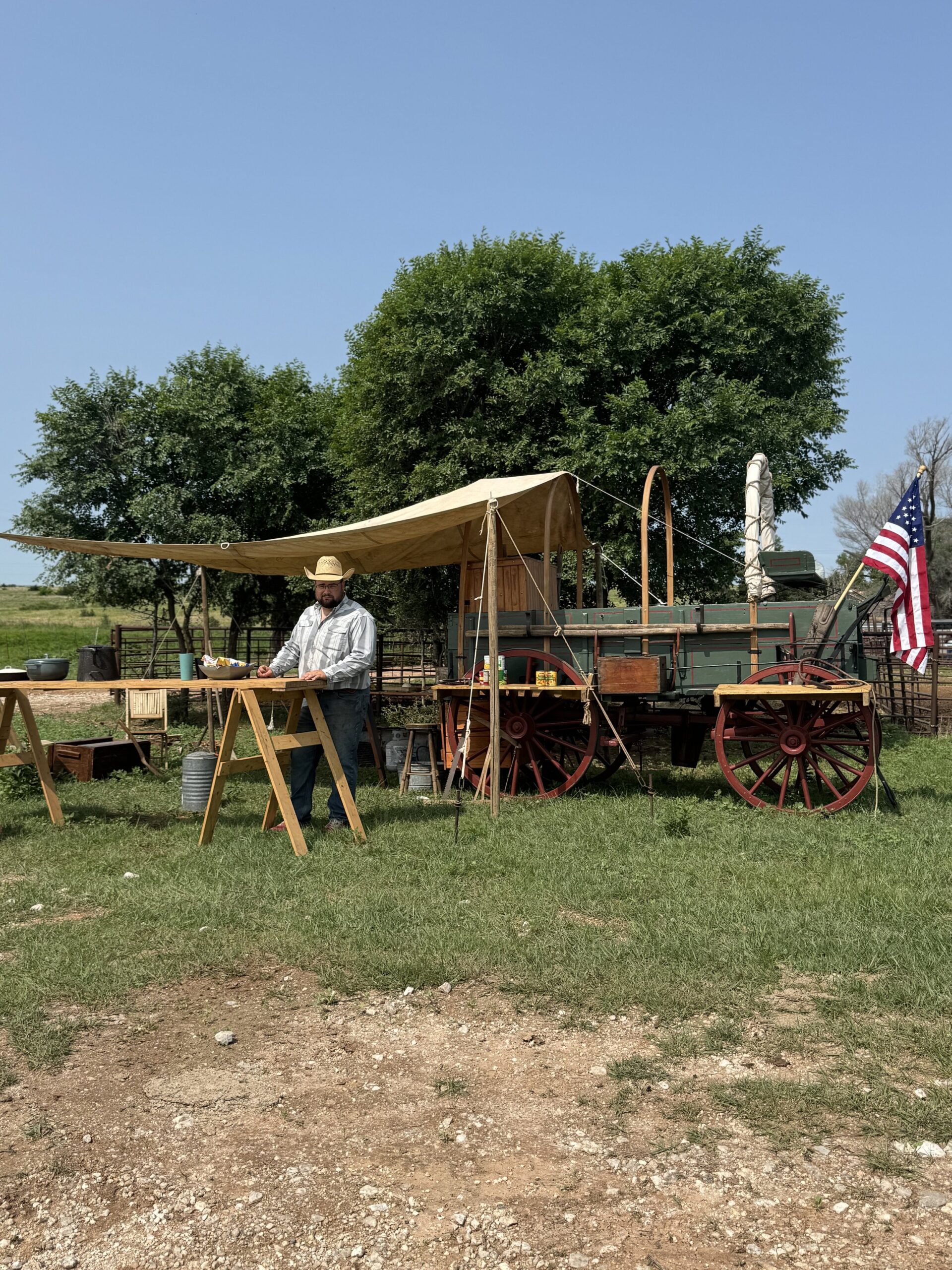 Man in a straw hat standing at wooden tables in front of a vintage wagon under a canopy, with trees and an American flag in the background on a sunny day.