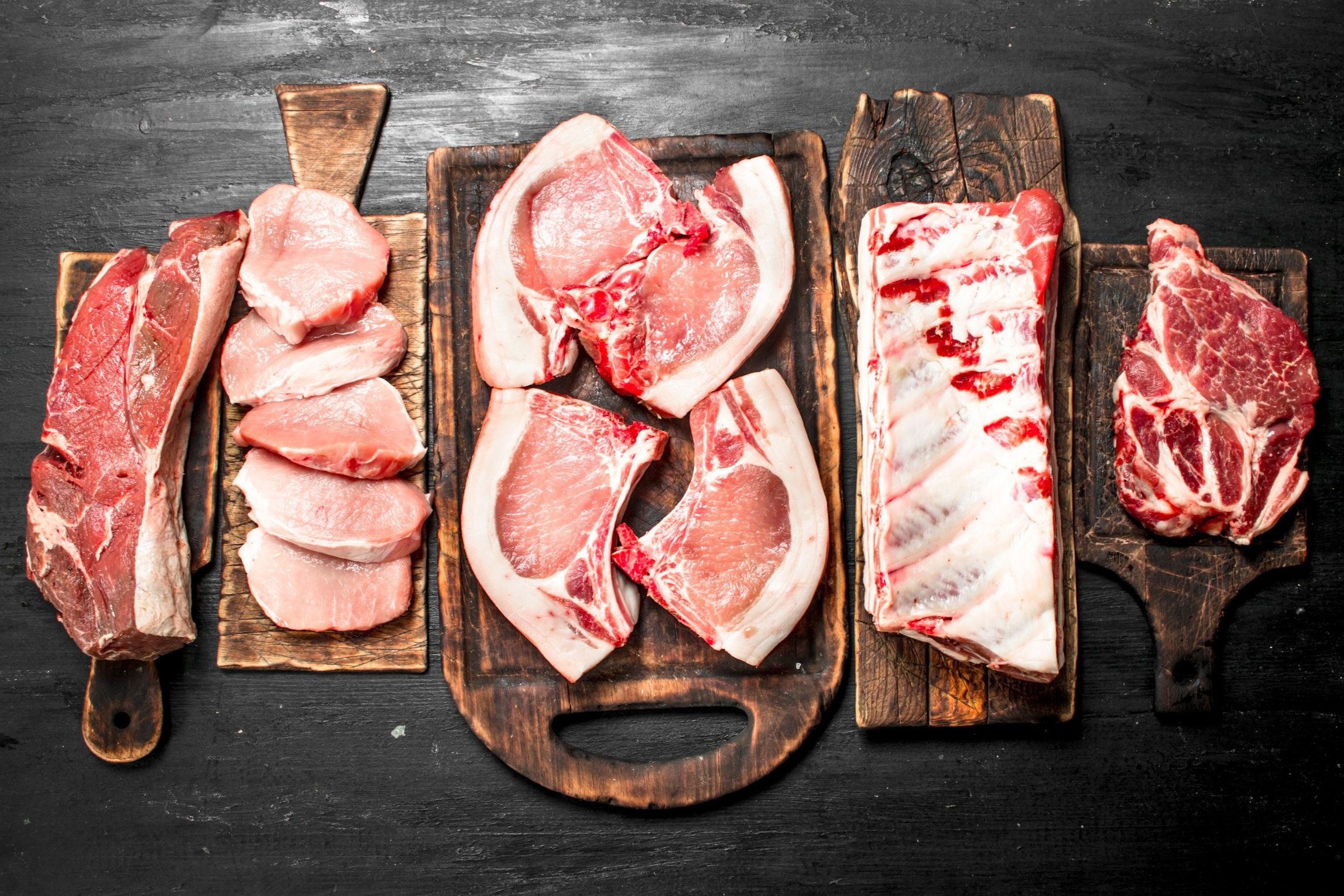 Assorted cuts of raw meat displayed on wooden cutting boards, including pork chops, rib racks, and beef steaks, against a dark background. Ideal for meat preparation and culinary presentations.