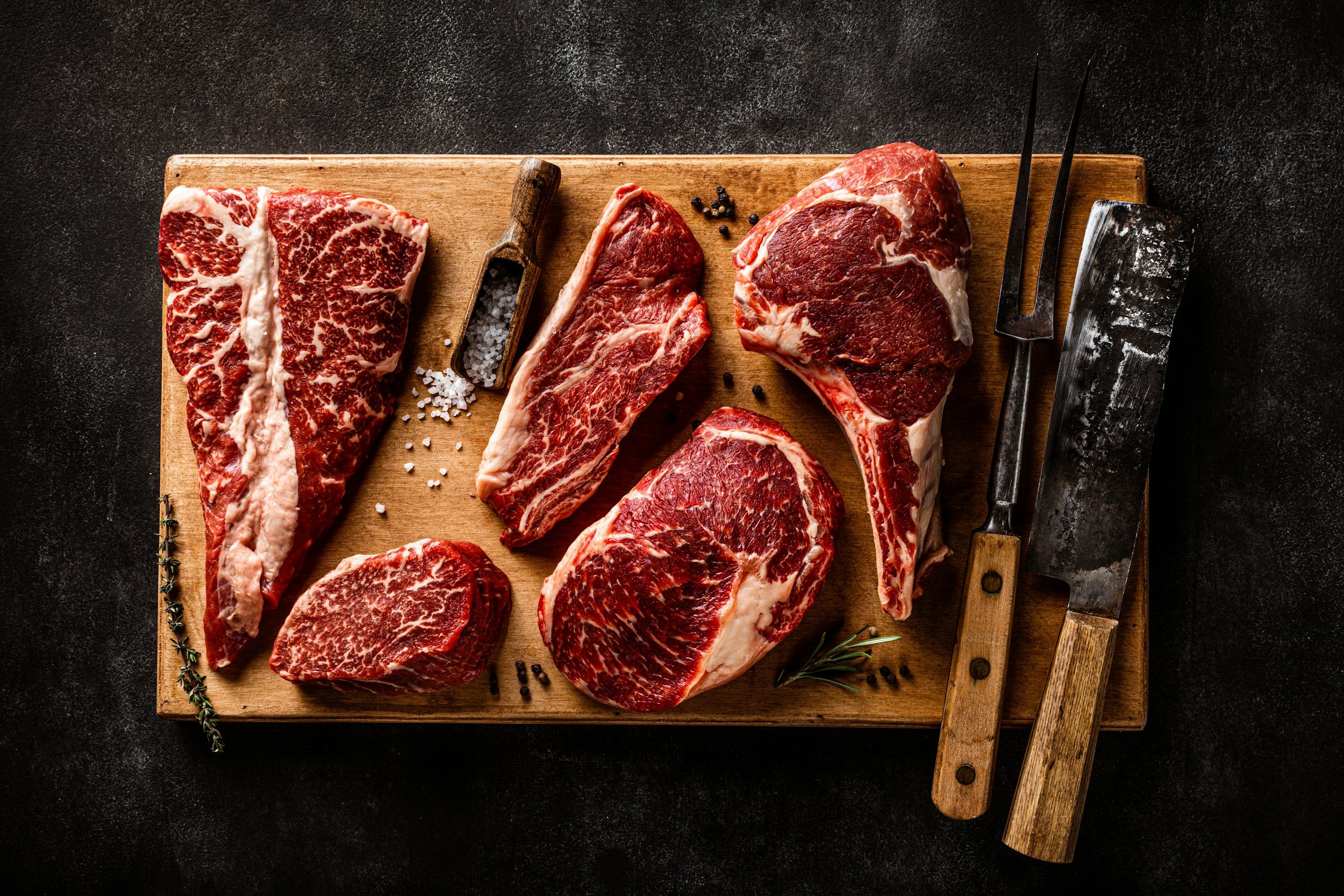 A selection of premium beef cuts arranged on a wooden cutting board, featuring marbled steaks, a butcher's knife, and seasoning like salt and pepper, set against a dark background.